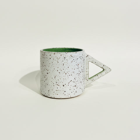 A speckled ceramic mug with a triangular handle, featuring a white exterior with dark brown and black speckles and a green interior, sitting on a wooden surface against a white wall.