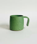 A green speckled ceramic mug with a thick handle, sitting on a wooden shelf against a plain off-white wall.