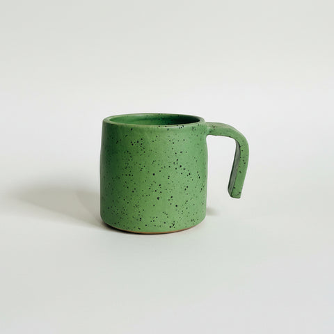 A green speckled ceramic mug with a thick handle, sitting on a wooden shelf against a plain off-white wall.