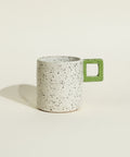 A speckled ceramic mug with a light gray body and a green square-handle accent, sitting on a wooden table against a white wall.