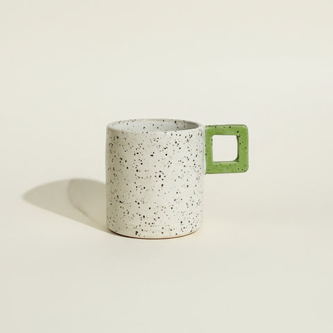 A speckled ceramic mug with a light gray body and a green square-handle accent, sitting on a wooden table against a white wall.