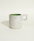A speckled ceramic mug with a square handle and a green interior, sitting on a wooden shelf against a plain wall.