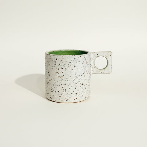 A speckled ceramic mug with a square handle and a green interior, sitting on a wooden shelf against a plain wall.