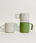 Three handmade ceramic mugs with unique handle shapes displayed on a wooden shelf against a white wall.