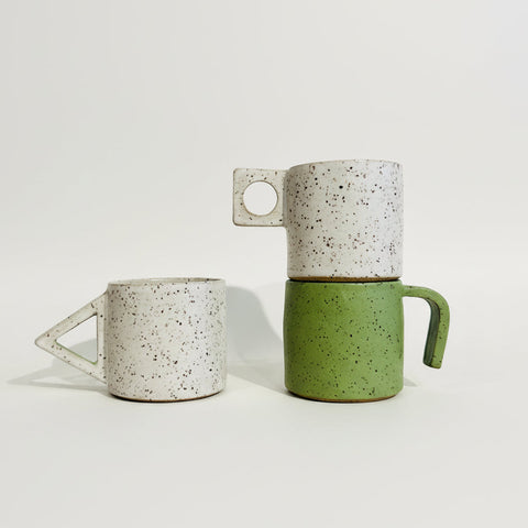 Three handmade ceramic mugs with unique handle shapes displayed on a wooden shelf against a white wall.