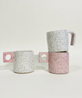 Two speckled ceramic mugs with unique handle designs, one standing alone on a wooden shelf and the other stacked on top of the other, set against a plain white wall.