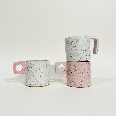 Two speckled ceramic mugs with unique handle designs, one standing alone on a wooden shelf and the other stacked on top of the other, set against a plain white wall.