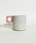 A handmade ceramic mug with a speckled off-white body and a pink handle featuring a circular cutout, sitting on a wooden surface against a plain white wall.