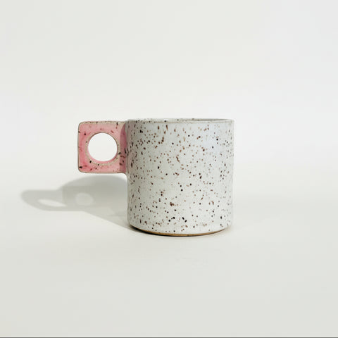 A handmade ceramic mug with a speckled off-white body and a pink handle featuring a circular cutout, sitting on a wooden surface against a plain white wall.