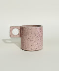 A pink speckled ceramic mug with a square handle featuring a circular cutout, resting on a wooden surface against a white wall.