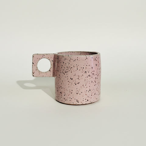 A pink speckled ceramic mug with a square handle featuring a circular cutout, resting on a wooden surface against a white wall.