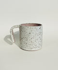 A handmade ceramic mug with a speckled off-white exterior and a dusty pink interior, sitting on a wooden shelf against a textured white wall.