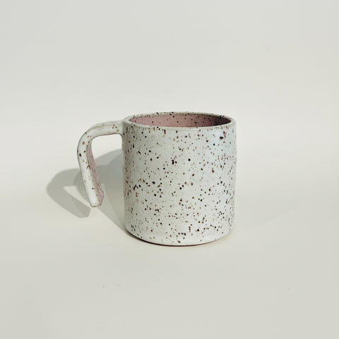 A handmade ceramic mug with a speckled off-white exterior and a dusty pink interior, sitting on a wooden shelf against a textured white wall.