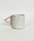 A speckled ceramic mug with a pink triangular handle, sitting on a wooden shelf against a plain off-white wall.