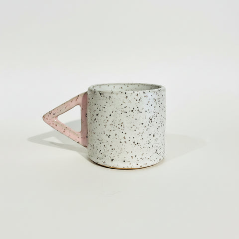 A speckled ceramic mug with a pink triangular handle, sitting on a wooden shelf against a plain off-white wall.