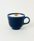 A dark blue ceramic mug filled with white wax, featuring a small wooden tag on top and a handwritten price tag hanging from the handle, set on a wooden table.