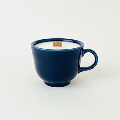 A dark blue ceramic mug filled with white wax, featuring a small wooden tag on top and a handwritten price tag hanging from the handle, set on a wooden table.