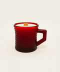 A textured dark red glass mug candle with white wax and a wooden wick sits on a wooden table, featuring a handwritten paper tag attached to the handle.