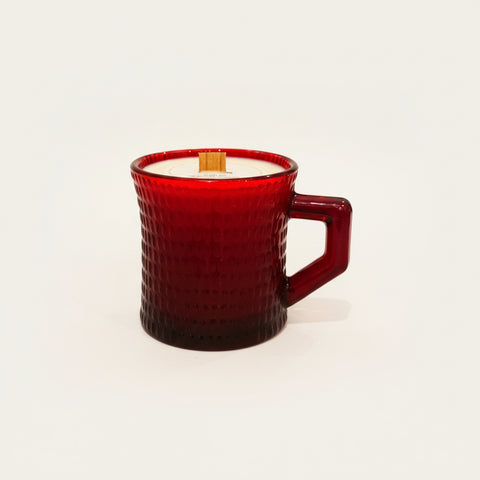 A textured dark red glass mug candle with white wax and a wooden wick sits on a wooden table, featuring a handwritten paper tag attached to the handle.