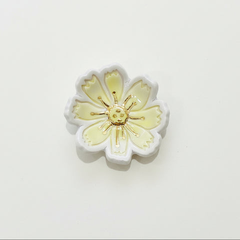 A small ceramic flower-shaped dish or jewelry stand with pale yellow and white petals and a gold-toned center, placed on a light wood surface.