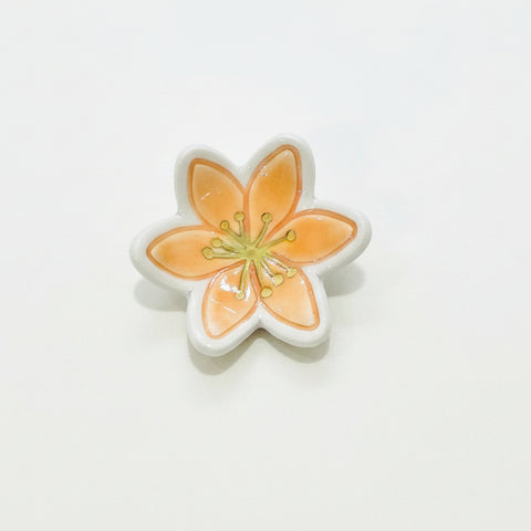 A small flower-shaped ceramic dish or trinket holder with orange petals, a white border, and a gold-toned center, placed on a light wood surface.