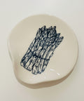 An irregularly shaped cream-colored ceramic dish featuring a black ink illustration of a bundle of asparagus, displayed on a light wood grain surface.