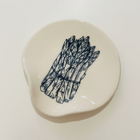 An irregularly shaped cream-colored ceramic dish featuring a black ink illustration of a bundle of asparagus, displayed on a light wood grain surface.