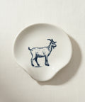 A small irregularly shaped cream-colored ceramic dish featuring a dark blue line-art illustration of a goat, placed on a light wood grain surface.