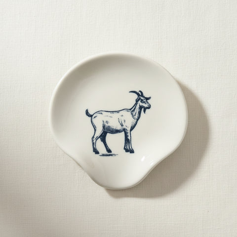 A small irregularly shaped cream-colored ceramic dish featuring a dark blue line-art illustration of a goat, placed on a light wood grain surface.