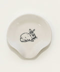 A small off-white ceramic dish with a black line drawing of a rabbit in the center, placed on a light wood tabletop.
