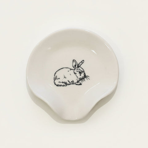 A small off-white ceramic dish with a black line drawing of a rabbit in the center, placed on a light wood tabletop.
