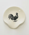 A small off-white ceramic dish with a black rooster illustration, placed on a light wood tabletop.
