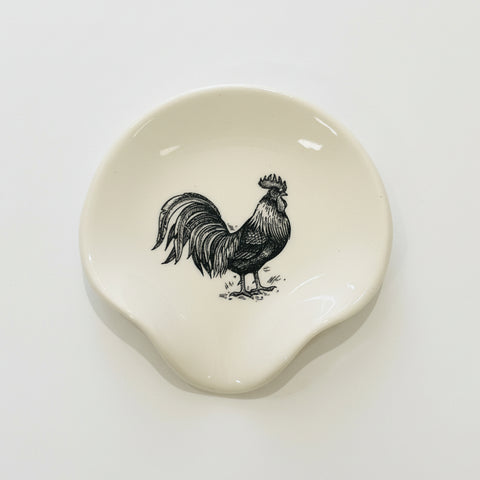 A small off-white ceramic dish with a black rooster illustration, placed on a light wood tabletop.