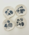 Four small white ceramic coasters with irregular organic shapes, decorated with dark blue mushroom illustrations, arranged on a light wood tabletop.