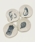 Four small white ceramic coasters with hand-drawn blue vegetable illustrations, arranged on a wooden tabletop.