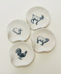 Four small white ceramic dish holders with irregular rounded shapes, each printed with a different animal illustration in dark blue: a goat, rabbit, sheep, and rooster. They are arranged on a light wood tabletop.