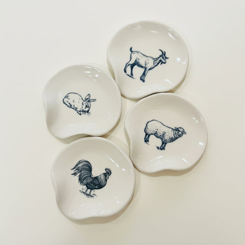 Four small white ceramic dish holders with irregular rounded shapes, each printed with a different animal illustration in dark blue: a goat, rabbit, sheep, and rooster. They are arranged on a light wood tabletop.