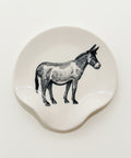 A round off-white ceramic coaster featuring a black line-art illustration of a donkey, placed on a light wood tabletop.