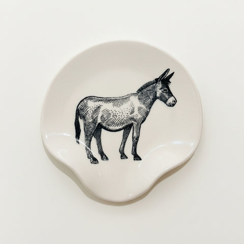 A round off-white ceramic coaster featuring a black line-art illustration of a donkey, placed on a light wood tabletop.