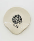 A small irregular-shaped cream ceramic dish with a black line-art illustration of a morepenny mushroom, placed on a light wood grain surface.