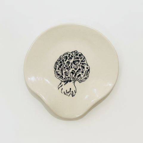 A small irregular-shaped cream ceramic dish with a black line-art illustration of a morepenny mushroom, placed on a light wood grain surface.