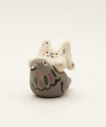 A small ceramic figurine of a gray bird with a white cat on its back, decorated with hand-painted black, pink, and blue details, sitting on a wooden table.
