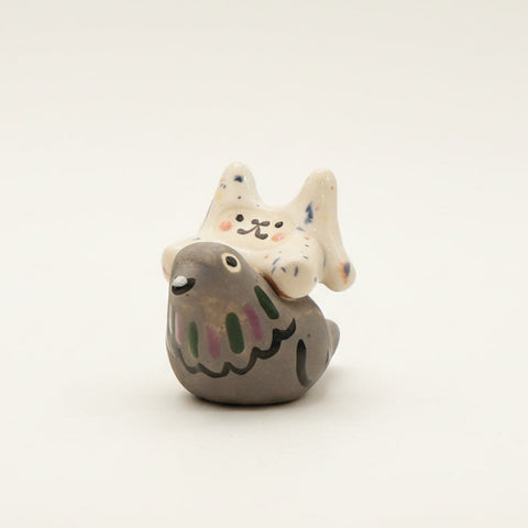 A small ceramic figurine of a gray bird with a white cat on its back, decorated with hand-painted black, pink, and blue details, sitting on a wooden table.