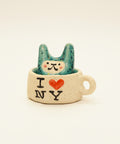 A ceramic rabbit-shaped cup with a teal speckled rabbit sitting inside, printed with 'I [heart] NY' on the side, placed on a wooden desk with other similar cups blurred in the background.