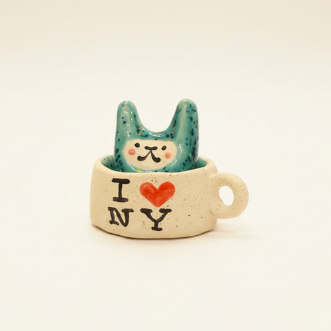 A ceramic rabbit-shaped cup with a teal speckled rabbit sitting inside, printed with 'I [heart] NY' on the side, placed on a wooden desk with other similar cups blurred in the background.