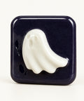 A square dark purple soap with a raised white ghost design on the front, wrapped in clear plastic and sitting on a wooden surface against a white wall.