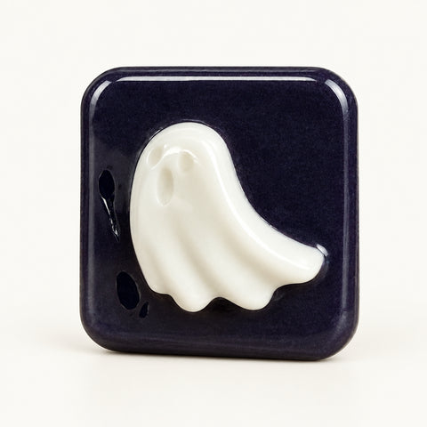 A square dark purple soap with a raised white ghost design on the front, wrapped in clear plastic and sitting on a wooden surface against a white wall.