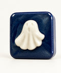 A square dark blue soap bar with a raised white ghost design on the front, photographed standing on a wooden surface against a plain light wall.