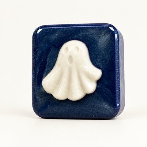 A square dark blue soap bar with a raised white ghost design on the front, photographed standing on a wooden surface against a plain light wall.