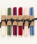 Six double taper candles in assorted colors wrapped in brown corrugated paper bands and tied with black ribbons, arranged on a light wood surface.